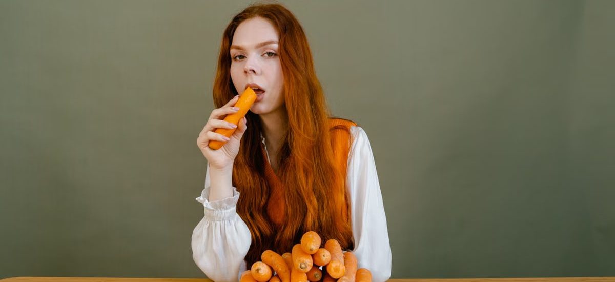 Red-haired woman sits at a table, holding a carrot, surrounded by a stack of vegetables.