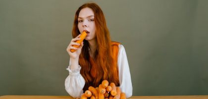 Red-haired woman sits at a table, holding a carrot, surrounded by a stack of vegetables.