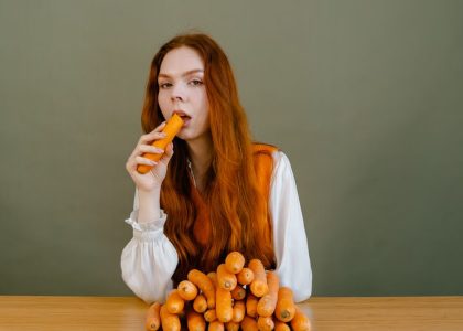 Red-haired woman sits at a table, holding a carrot, surrounded by a stack of vegetables.